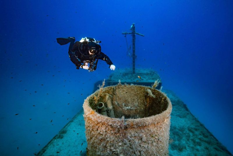 Un sub si avvicina alla parte superiore di un relitto. È circondato da pesciolini che nuotano nelle acque trasparenti.