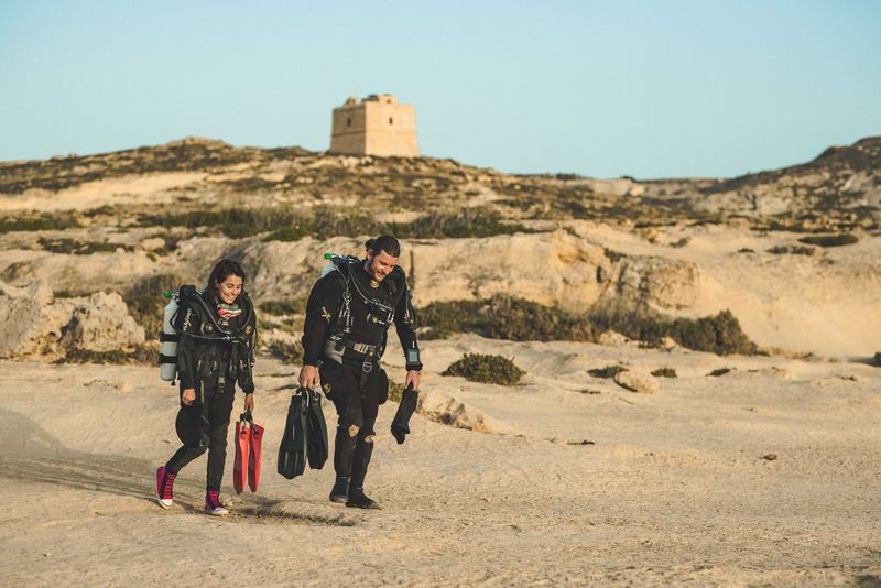 Due figure in muta da sub camminano nella sabbia, su uno sfondo di dune caratterizzato da una costruzione beige.