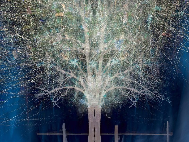 A tree with pale brown wood and green and gold foliage stands against a navy blue background. The colours are flat and artificial, while dots of moving lights surround the tree.