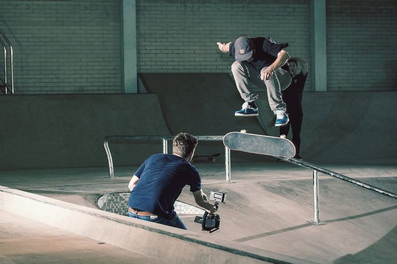 A skateboarder performs a jump by a rail in an indoor skate park, while a man kneels down in front of him holding out a video camera to capture the action.