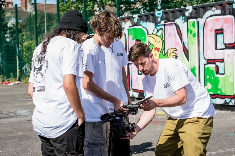 A group of people in white t-shirts stand outside in a sunny concrete park, gathered around a video camera and looking at the fold-out screen.