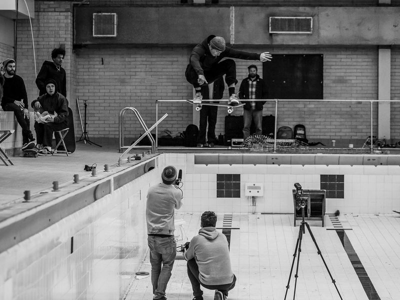 A black and white photo of a skateboarder performing a jump while being filmed by two men standing below and to the side of him.