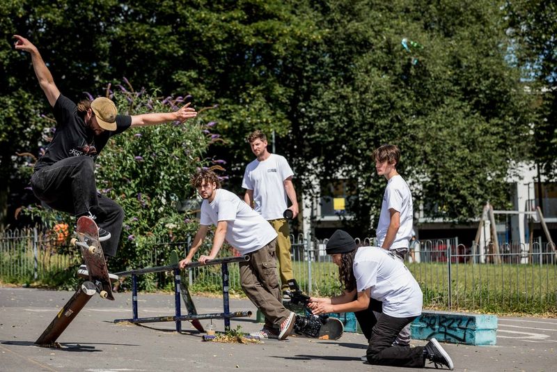 A skateboarder performs a jumper in an outdoor park, watched by four people in white t-shirts, one of whom is kneeling down and filming him on a video camera.
