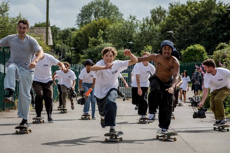 A large number of people on skateboards are skating towards the camera, looks of excitement and determination on their faces. A few of them are holding video cameras at calf level to film the action.