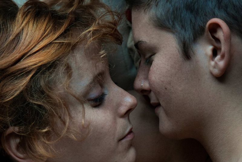 A close-up of two young people, one with red hair, one with short brown hair, with their noses almost touching. 