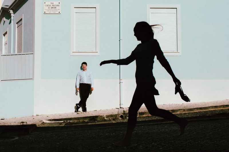 In una fotografia scattata da Alison Bounce con Canon EOS R5, un uomo vestito elegante si appoggia con disinvoltura al muro di una casa, mentre accanto a lui si staglia la sagoma molto più grande di una donna che cammina portando in mano un paio di scarpe col tacco.