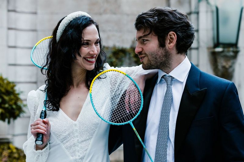 A bride and groom each hold a badminton racket as they smile playfully at each other in a photograph taken by Alison Bounce.