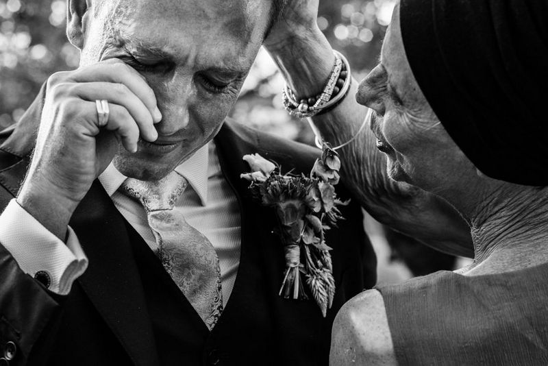 A man in a suit wipes a tear from his eye as a woman smiles and places a hand to his head, in a black and white wedding photograph by Alison Bounce.
