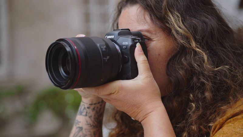 Photographer Alison Bounce holds a Canon EOS R5 camera up to her face, looking through the viewfinder.