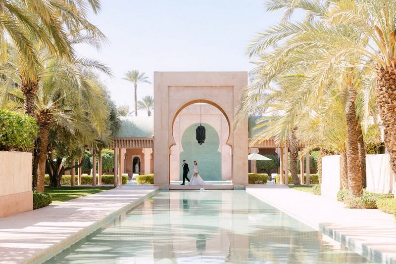 A panoramic shot of a wedding couple walking under a moorish arch, with a pool of water flanked by palm trees leading the eye to them. Location wedding photo by Rebecca Carpenter.