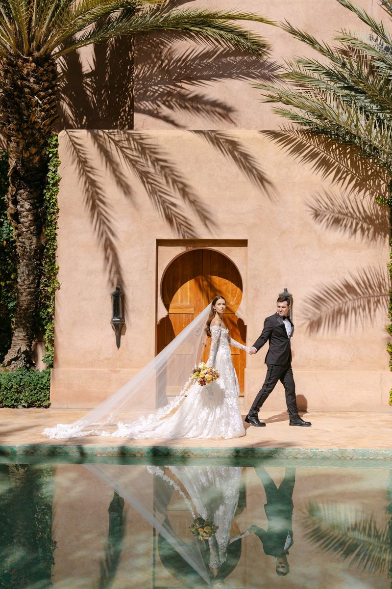 A bride and groom stroll past a moorish-arch door, with a pool of water reflecting them below and the shadows of palm trees on the stone wall behind. Location wedding photo by Rebecca Carpenter