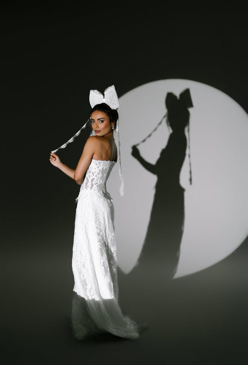 A bride wearing an off-the-shoulder wedding gown and a large bow on her head poses for the camera, a sharp shadow behind thrown by a round spotlight. Wedding photo by Rebecca Carpenter.