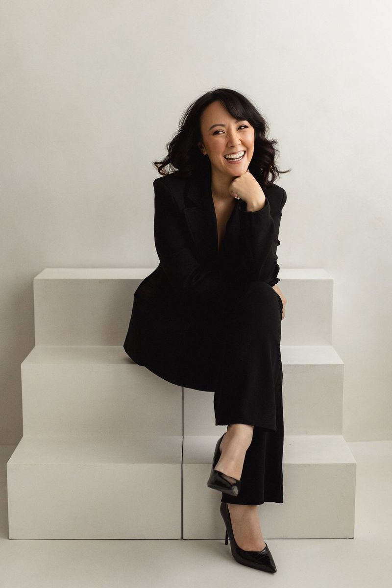Wedding photographer and content creator Rebecca Carpenter, dressed in a black suit, sits on a set of white prop steps, smiling at the camera.