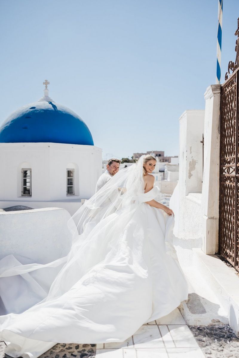 A wedding couple on a sunny rooftop, the bride’s dress billowing behind her. A blue dome with a cross on it and closely-packed rooftops are visible in the background. Location wedding photo by Rebecca Carpenter.
