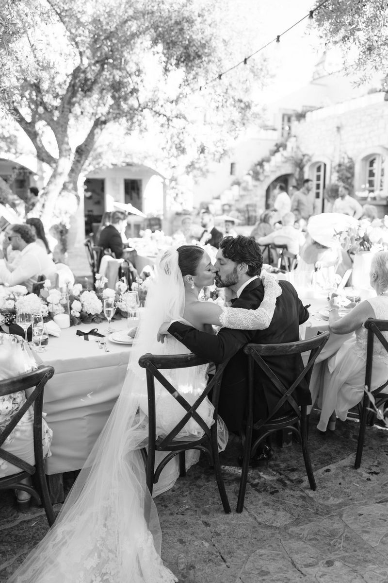 A black and white photo of a bride and groom kissing, seated at a wedding banquet in a courtyard in bright daylight with guests visible at the other tables. Location wedding photo by Rebecca Carpenter.