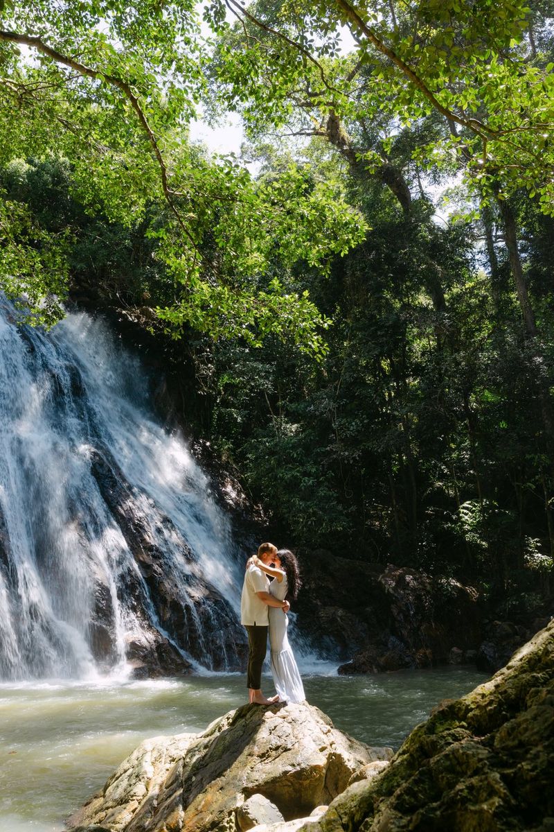 A couple embracing on a rocky outcrop with a waterfall and lush foliage in the background behind a pool of water. Location wedding photo by Rebecca Carpenter.