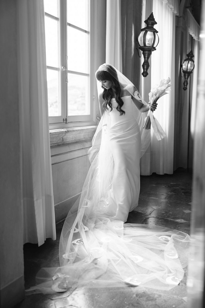 Black and white photo taken by Rebecca Carpenter showing a bride adjusting the train of her wedding dress in front of a window in a corridor with large wall lights behind.