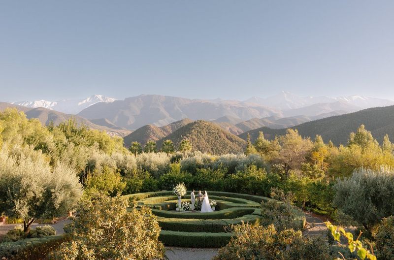 A panoramic location wedding photo by Rebecca Carpenter, showing a couple and celebrant standing in the centre of a low formal maze with low trees and bushes around them and a sweeping vista of mountains in the background.