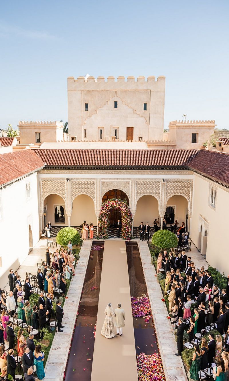 An aerial view of a wedding couple walking up the centre of a courtyard with a rill of water on either side and wedding guests to the edges. Location wedding photo by Rebecca Carpenter.