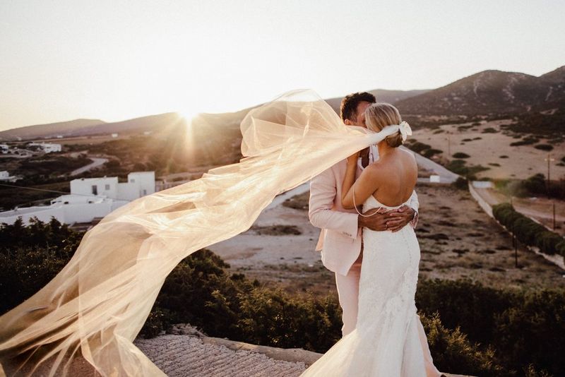Novelli sposi che si abbracciano al tramonto, il velo della sposa è mosso dalla brezza.