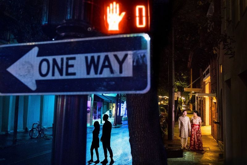 Sagoma di una coppia sotto un cartello di senso unico; un'altra coppia cammina sul marciapiede, illuminata dalle luci della strada.