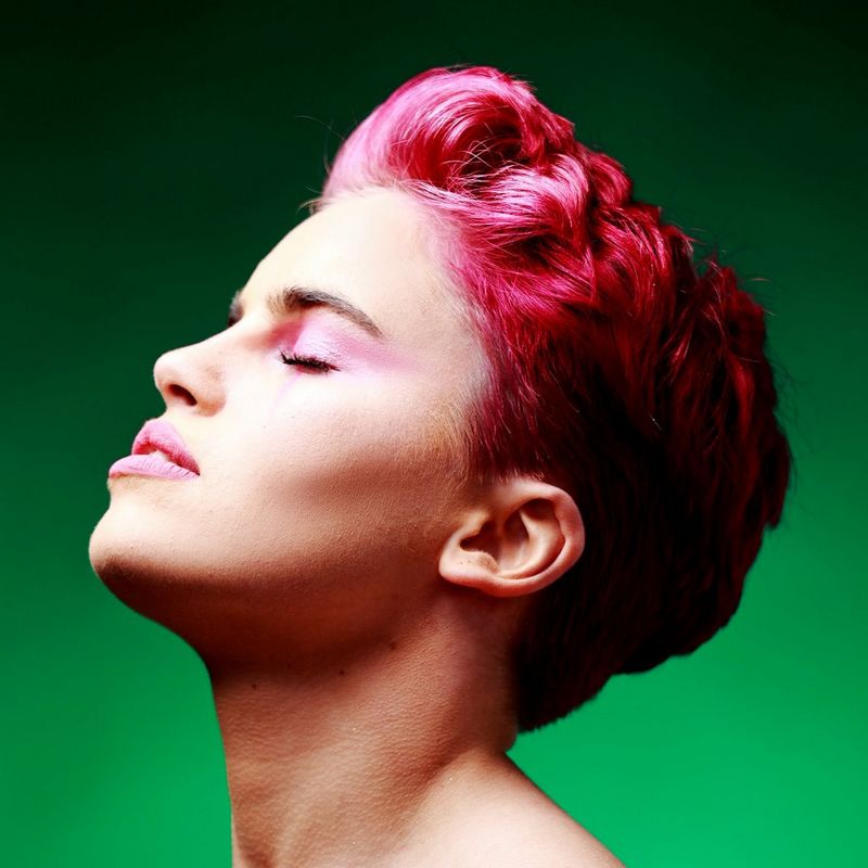 A photo taken by James Musselwhite of a person with pink hair and pink eye shadow and lip colour against a green background, closing their eyes and lifting their head.