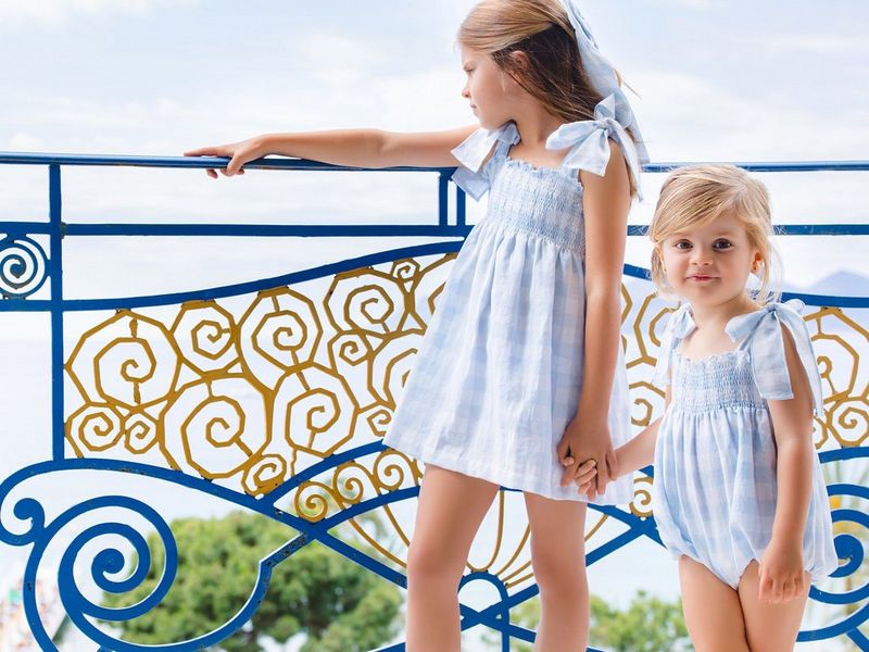 A photo taken by Marisa Martins showing two children in matching blue checked outfits holding hands as the one on the left places her arm on a dark blue railing with an ornate pattern inset. 