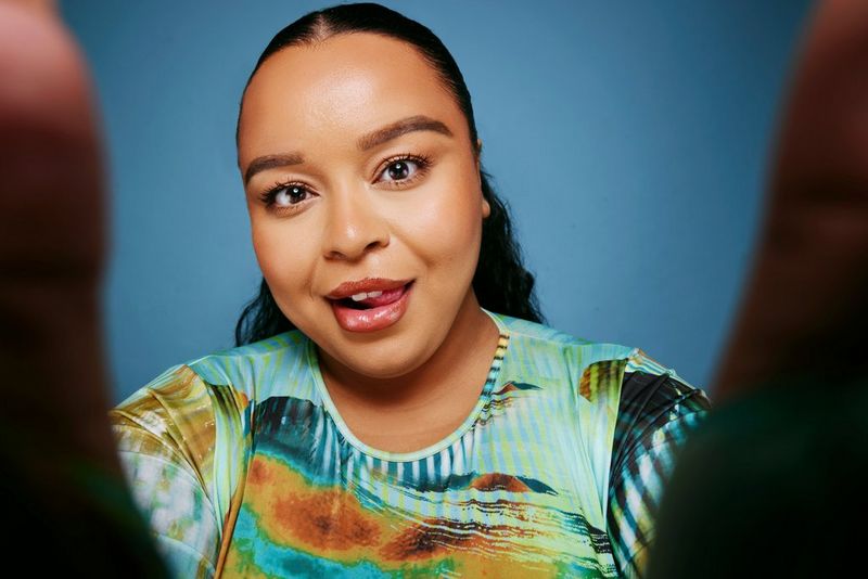 A photo taken by Jade Keshia Gordon of a woman pressing her tongue to the side of her mouth as she looks directly at the camera while her fingers frame the sides of the lens. 