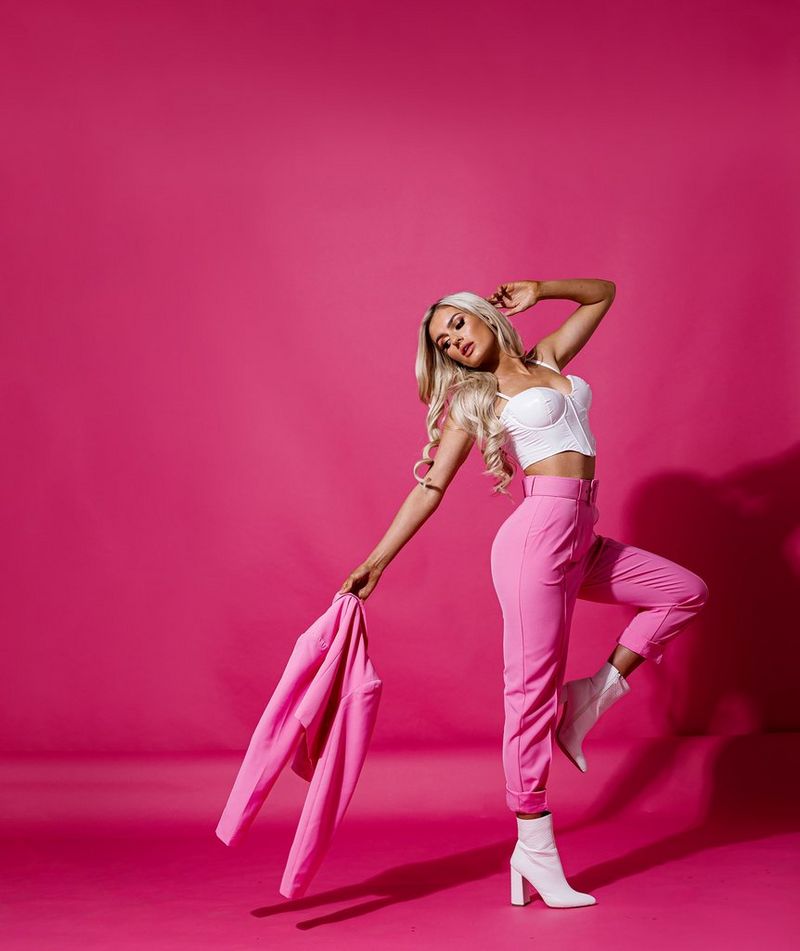 A photo taken by James Musselwhite showing a woman in pink trousers and a white top holding a pink jacket in her right hand and posing against a pink background. 