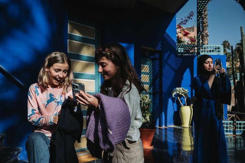 Against dark blue walls, two girls smile while looking at the screen of a smartphone while another woman to the right holds her smartphone up. Photo by Marisa Martins.