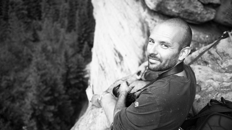 A black and white image of Jakub Frey sitting on the edge of a cliff face, looking back towards the camera. 