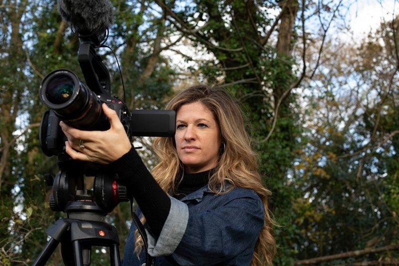 Une femme aux cheveux blonds foncés règle son appareil Canon alors qu'elle filme dans une forêt.