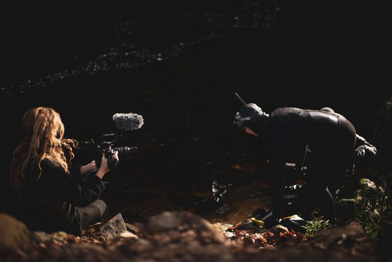 Un homme vêtu d'une tenue de plongée plonge dans une rivière tandis qu'une femme munie d'un appareil Canon le filme depuis la rive.