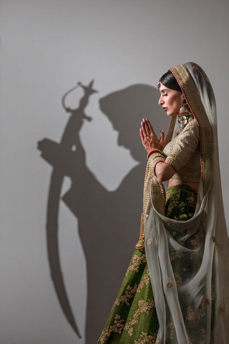 Zijaanzicht van een vrouw met een sluier in een lange, sierlijke jurk, de handen gevouwen in gebed. Op de muur achter haar is de schaduw te zien van een man in een soortgelijke houding, met een kromzwaard tussen de handen. Gemaakt door fotograaf Sanjay Jogia.
