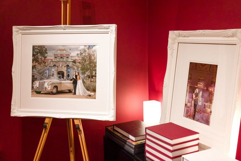 Op een ezel staat een grote ingelijste print van een trouwfoto waarop een bruid en bruidegom naast een crèmekleurige cabrio voor een groot gebouw poseren. Naast de ezel in een rode kamer ligt een stapel albums en staat nog een ingelijste print.