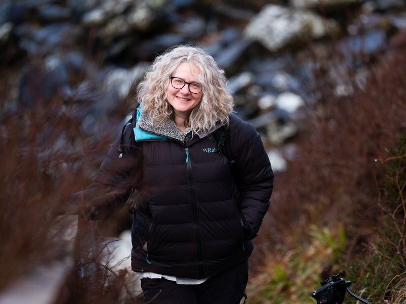 Filmmaker Heather Hughes lacht naar de camera, met de natuur op de achtergrond.