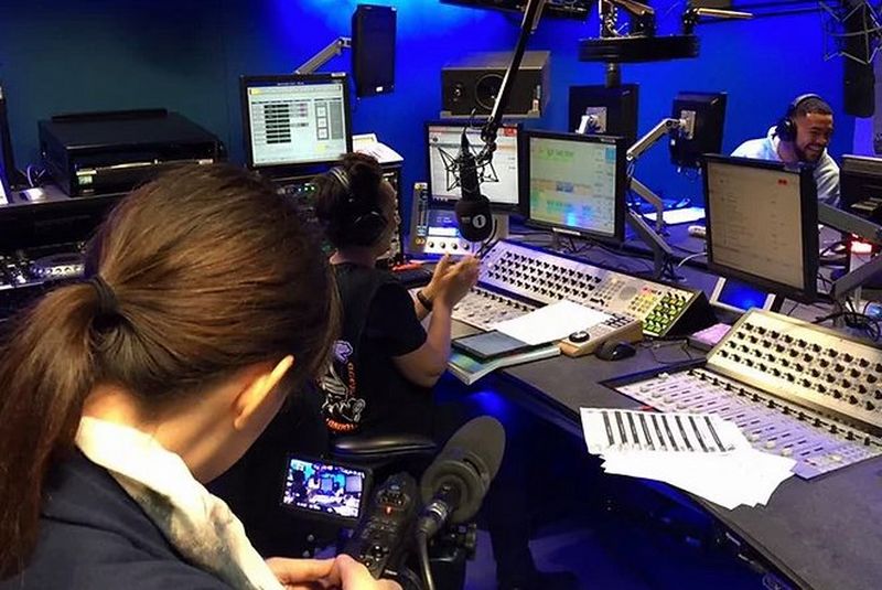 Een over-de-schouder-opname van filmmaker Carys Kaiser, die een vrouw aan een broadcasting desk filmt.