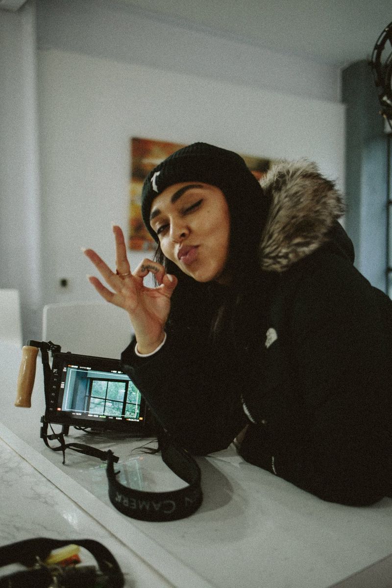 Regisseur Ashleigh Jadee lacht naar de camera en maakt het handgebaar voor 'oké', met haar camera in beeld.