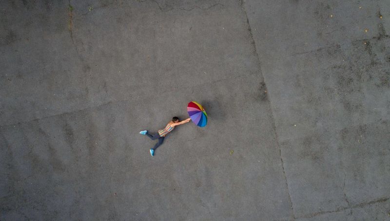 Een vrouw ligt op de grond en houdt een paraplu met regenboogkleuren vast, vastgelegd met een drone hoog in de lucht.