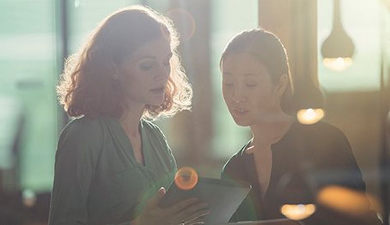 Two women, one with shoulder length curly red hair and the other with straight dark hair pulled back, look at a tablet screen.