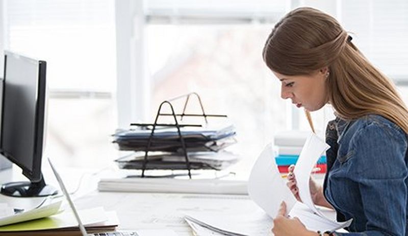 Woman with long brown hair sits at a computer desk covered with papers, looking through pages of a report.