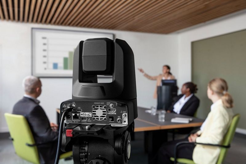 A Canon CR-N100 PTZ camera in a meeting room, recording a presenter pointing to a chart on the screen in the background.