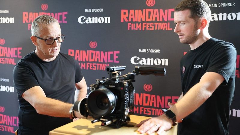 Deux hommes examinent une caméra de cinéma Canon devant une toile de fond du festival de cinéma Raindance portant l'inscription « Sponsor principal Canon ».