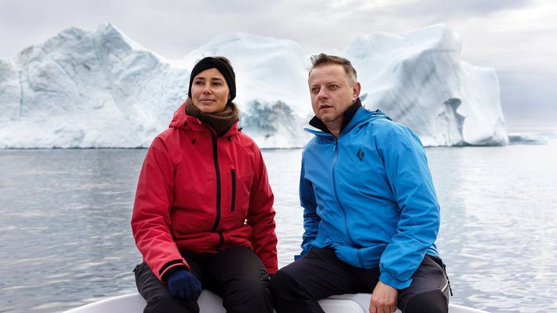 Tuperna Davidsen (left) in a red winter jacket, sits next to Rareș Beșliu, who wears a similar blue jacket. They are on the edge of a boat, looking beyond the camera at something unknown in the distance. Behind them is a huge white glacier.
