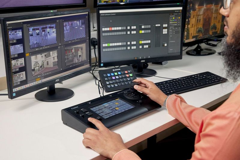 A user with a Canon RC-IP100 remote camera controller and a screen monitoring multiple camera feeds.