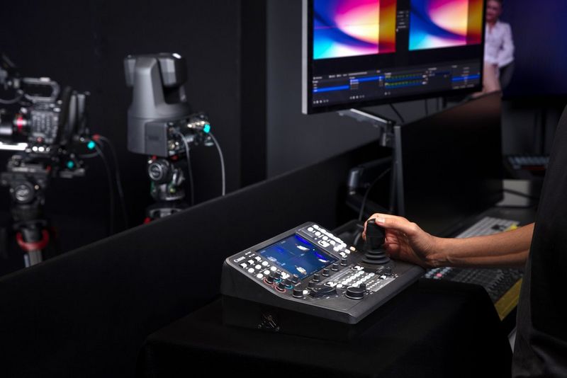 A user with a Canon RC-IP1000 PTZ camera controller in a dark studio, with a monitor screen and PTZ camera visible in the background.
