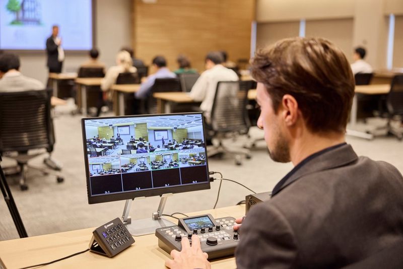 An operator using a Canon CANON RC-IP300 PTZ controller at the back of a classroom, with an external screen monitoring multiple camera feeds.
