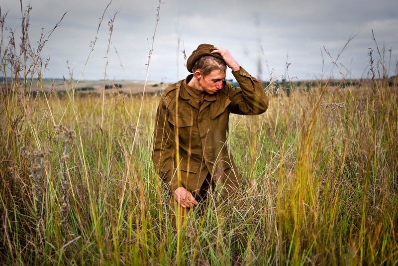 Африканский подросток с окровавленным лицом удрученно стоит, прижав одну руку к голове, в поле с высокой травой.