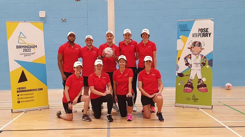 A netball team, posed for the camera with five people stood at the back and four on one knee at the front. They all wear red Canon branded polo shirts and white Canon branded baseball caps. The man stood at centre back holds a netball. On either side of them are pop up banners advertising the Birmingham 2022 Commonwealth Games.
