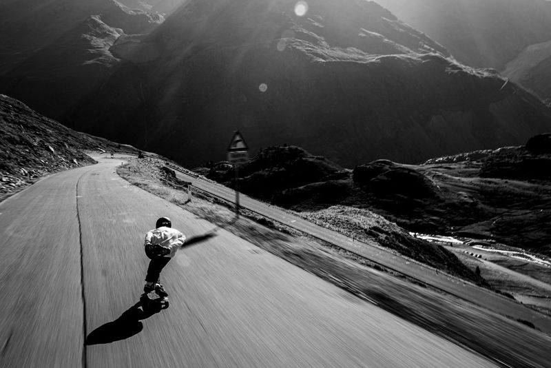 In a photo taken by Felix Pirker, a skateboarder viewed from behind speeds down a steep mountain road, with rugged alpine scenery in the background.